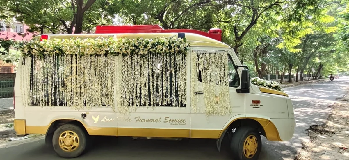 Last Ride Funeral premium glass hearse van with jasmine garland side curtains and floral crown moving along a tree-lined Delhi road during an Antim Yatra procession