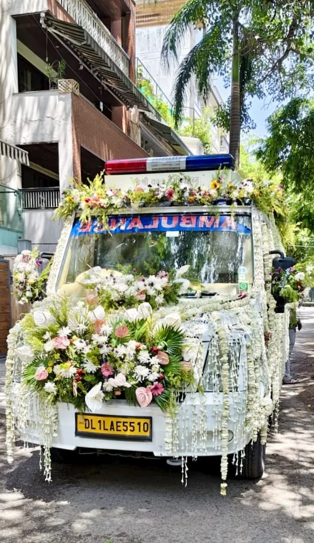 Front view of Last Ride Funeral premium hearse van with dense arrangement of white chrysanthemums, pink anthuriums, roses and jasmine garlands cascading over the bonnet — Delhi residential pickup for Antim Yatra